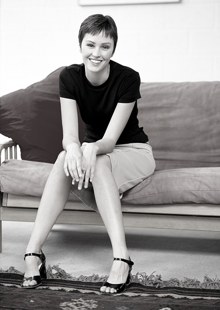 beautiful woman short brown hair wearing black shirt tan skirt funky shoes sits on a couch smiling